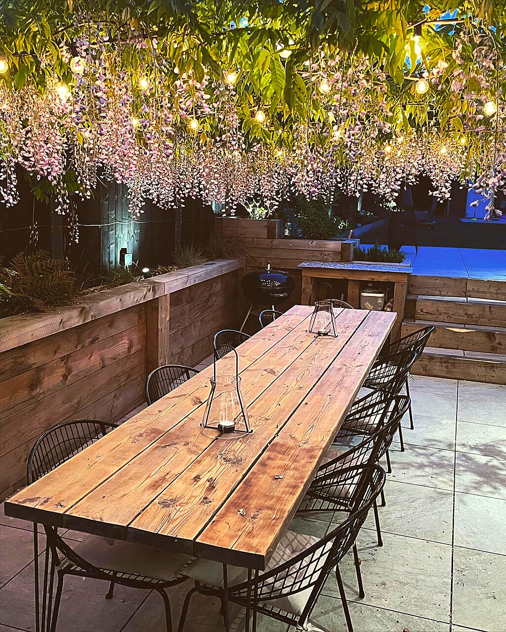 Garden terrace at dusk, with a long timber table under wisteria and festoon lights
