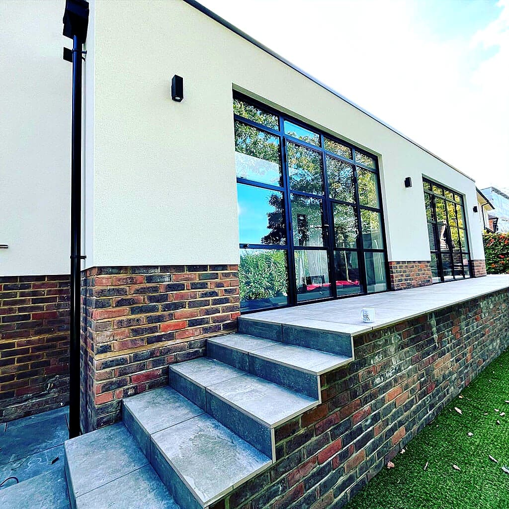 Single storey extension with black Crittall style glazing, cream render over a brick plinth, and stone steps up to a raised terrace
