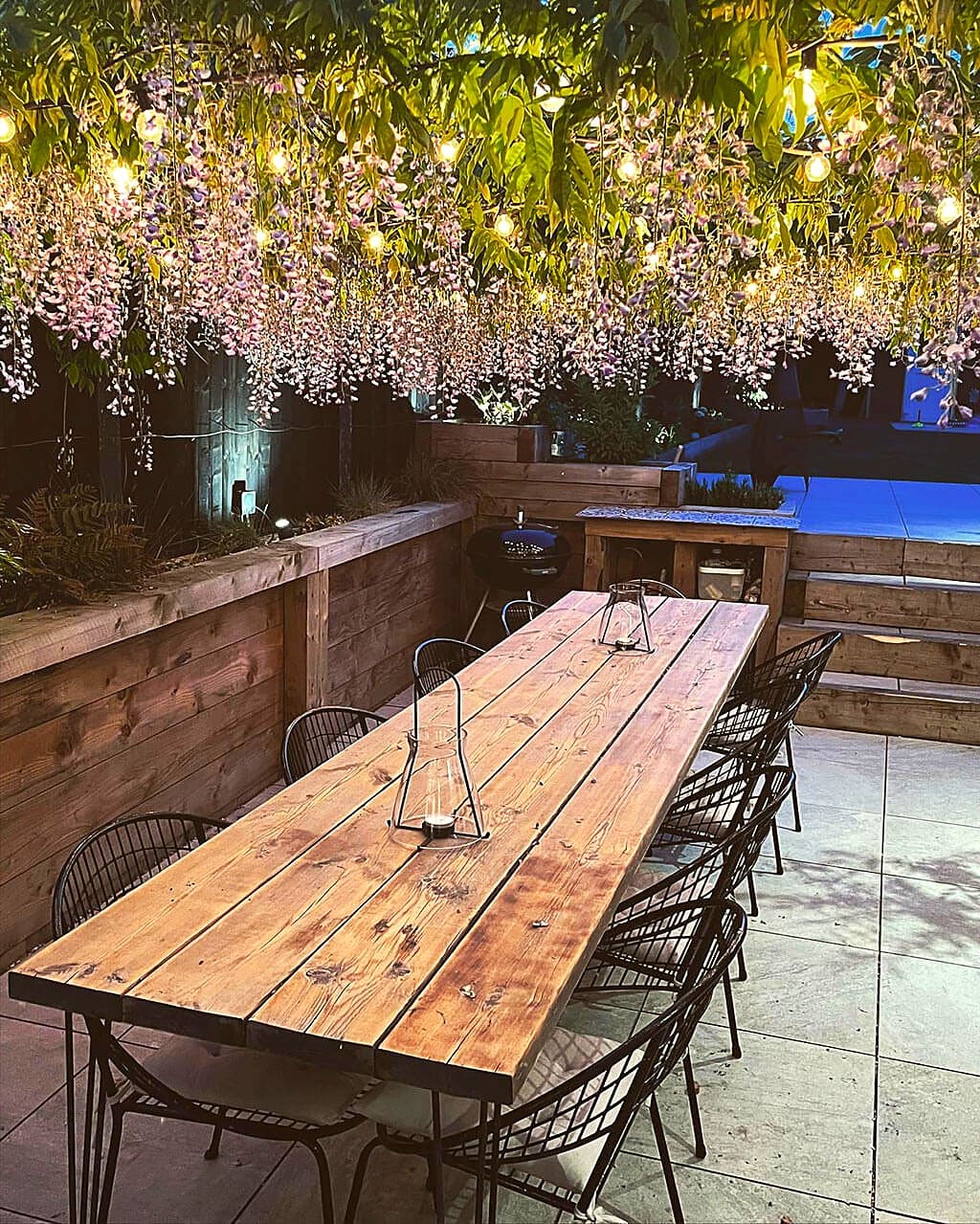 Garden terrace at dusk, with a long timber table under wisteria and festoon lights